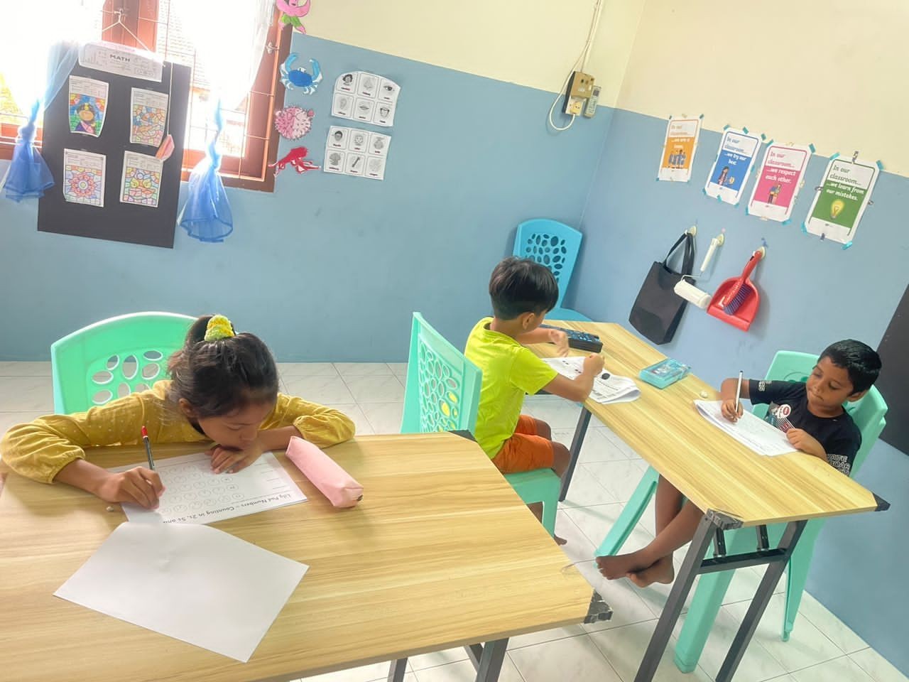 Three young children sitting at separate tables in a classroom, concentrating on their worksheets.