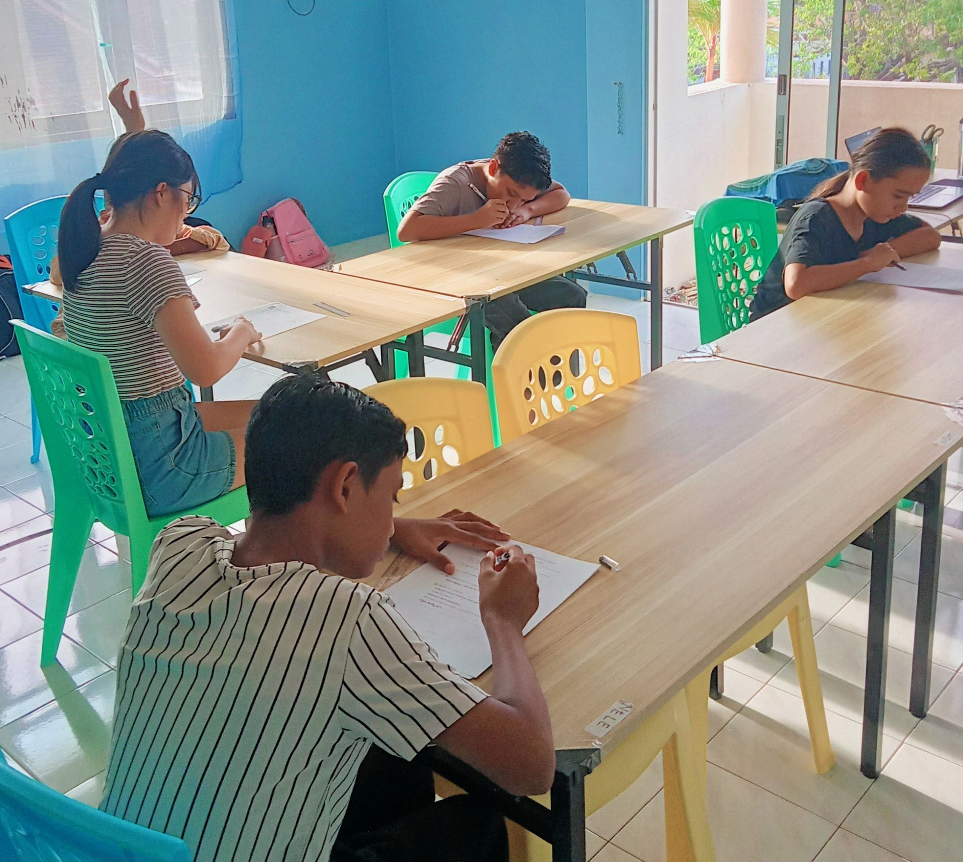 Students sitting at tables in a classroom, focusing on writing assignments with papers and pens.